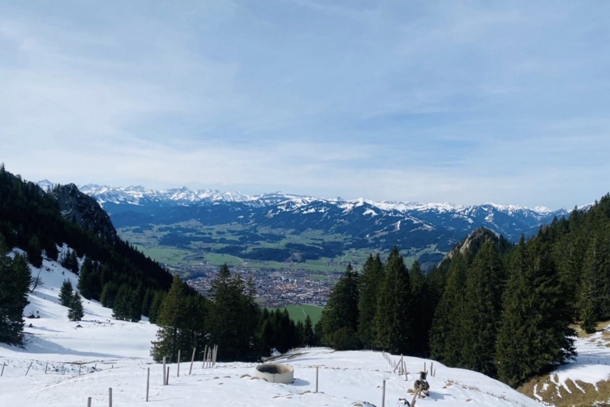 Blick vom Bergrücken hinunter auf Oberstdorf im Tal, Spätwinter mit teilweise schneebedeckten Wiesen, Allgäuer Alpen am Horizont