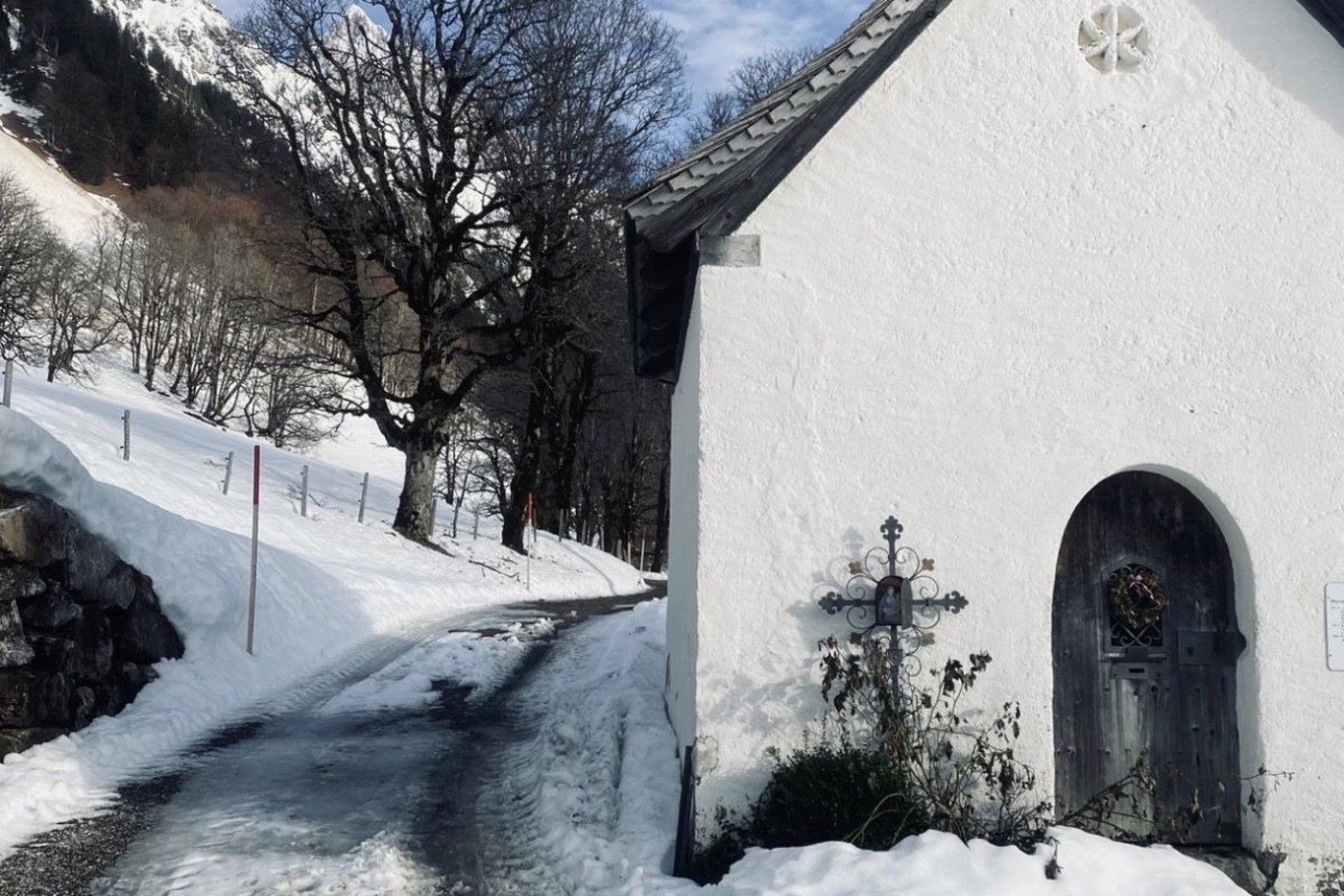 Whitewashed mountain chapel with a wrought-iron wayside cross and bare sycamore maple on a late-winter day