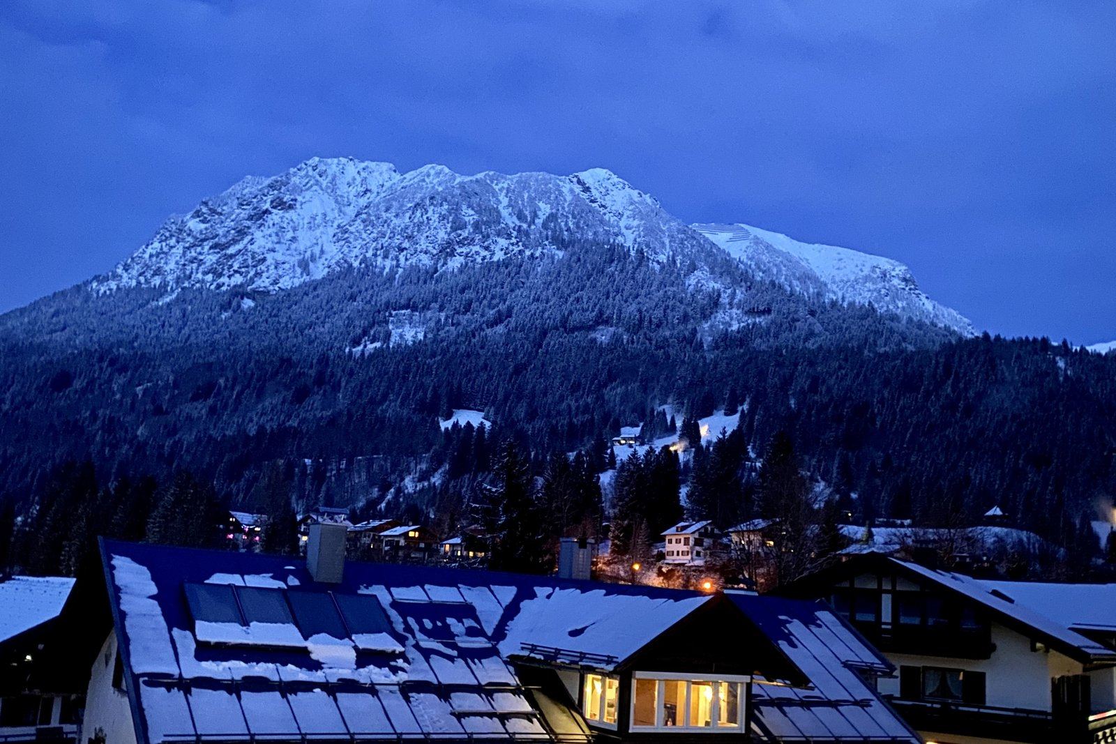 Blaue Stunde im Winter, schneebedeckter Berg im letzten Tageslicht, warm beleuchtete Fenster der Häuser im Vordergrund