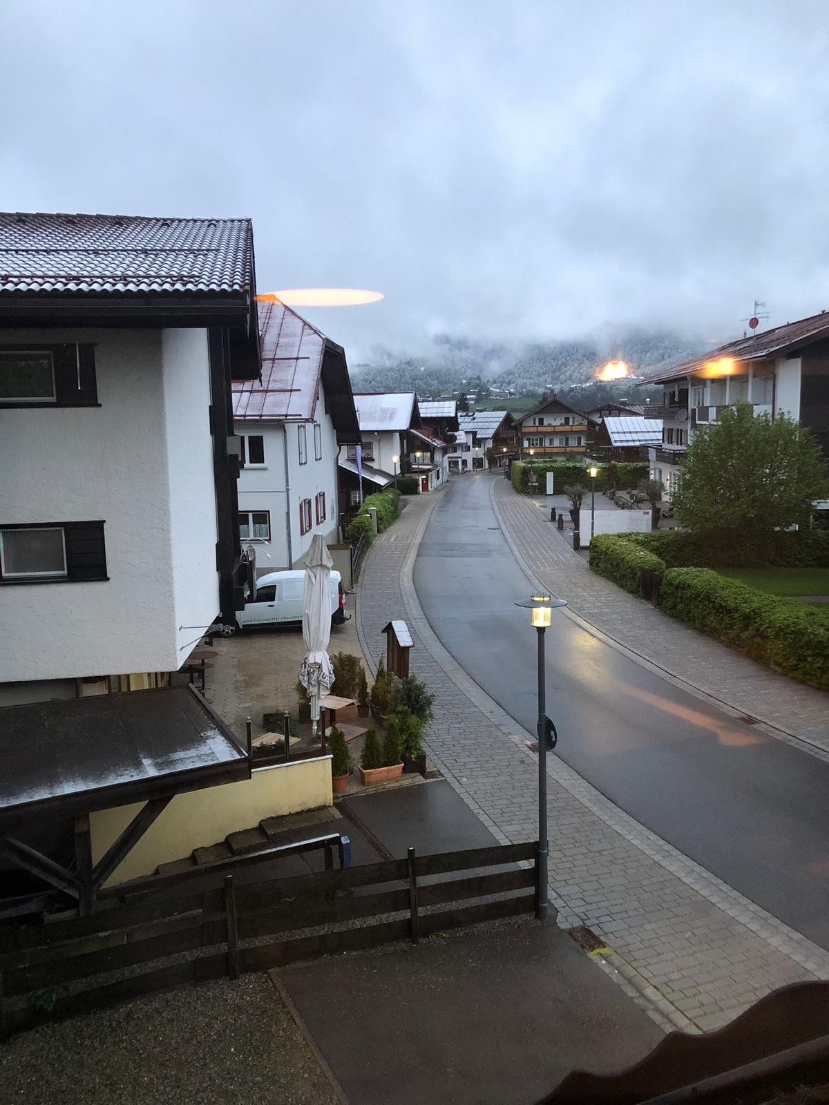 Regenmorgen, leere Pflasterstraße, Straßenlaternen brennen, Wolken hängen tief über den Allgäuer Bergen